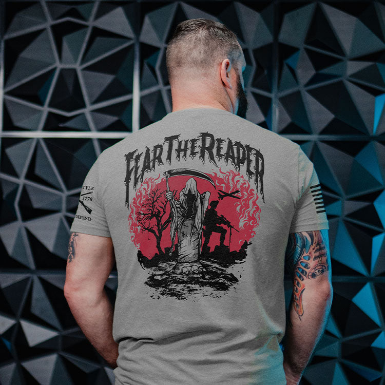 A man viewed from behind wearing a heather gray t-shirt with a large black and red graphic on the back that reads 'FEAR THE REAPER'. The background consists of dark, geometric, 3D-walled paneling.